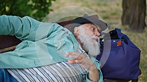 Portrait of senior sleeping homeless man waking up sitting down on bench in park looking away. Sad depressed helpless