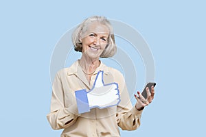 Portrait of senior woman with cell phone holding fake like button against blue background