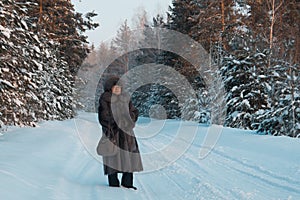 Portrait of senior woman fur coat and hat standing in cold winter snow covered forest, telephoto