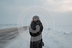 Portrait of senior woman fur coat and hat standing in cold winter snow covered road, telephoto