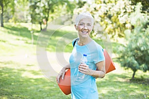 Senior woman holds fitness mat on her back in the park and preparing for exercise