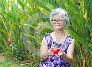 Portrait of a senior woman with short gray hair, wearing glasses, holding a red heart shape, smiling, and looking at the camera