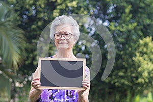 Portrait of a senior woman with short gray hair, wearing glasses, smiling, holding a mini blackboard, and looking at the camera