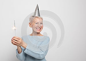 Portrait of a senior woman in studio on a gray background. Woman wearing blue sweater and party hat, holding sparkles.
