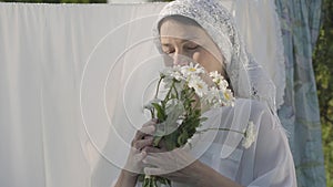 Portrait senior woman with white shawl on her head sniffing daisies looking at camera near the clothesline outdoors