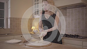 Portrait sensual blond woman with big breasts in black dress knead the dough with sigaret in mouth on the kitchen table