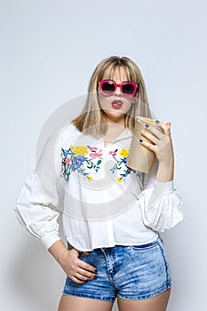 Portrait of Sensual Caucasian Female Blond Girl In Shorts and Red Sunglasses Holding Popcorn Bucket and Posing Against White