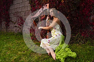 Portrait of Sensual caucasian female harpist in long decorated dress posing with harp against ivy wall outdoor