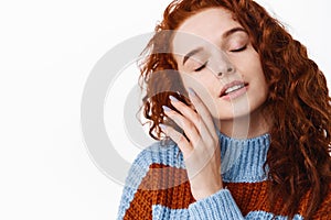 Portrait of sensual redhead woman with natural curly ginger hair, touching her smooth clean facial skin with closed eyes