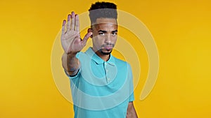 Portrait of serious african american man showing rejecting gesture by stop palm sign. Guy isolated on yellow background.