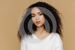 Portrait of serious beautiful dark skinned female with frizzy black hair, has minimal makeup, looks calmly at camera, wears white