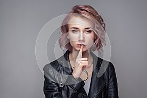 Portrait of serious beautiful girl with short hairstyle and makeup in casual leather jacket standing with silent finger on lips