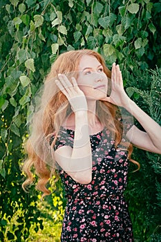 Portrait of a serious beautiful redhead lady in summer dress fixing her hair with hands under birch tree in spring time