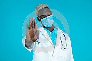 Portrait of serious black doctor in professional medical white coat and mask showing rejecting gesture by stop palm sign