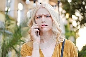 Portrait of serious blond girl biting lip and scaredly looking in camera while talking on cellphone outdoor