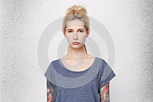 Portrait of serious blonde girl wearing blue t-shirt, tattoed arms and pierced nose sad and confused. She lost her boyfriend