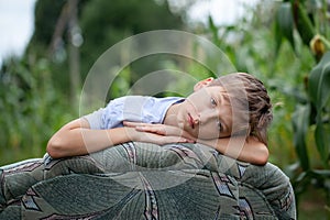Portrait serious boy sitting on a armchair in a field in the countryside in summer day