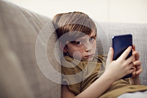 Portrait Of Serious Boy Sitting On Sofa At Home Gaming On Hand Held Devices At Home