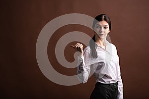 Portrait of serious brunette business woman pointing her thumb to the side in studio on brown background with copyspace.