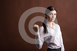 Portrait of serious brunette business woman pointing her thumb to the side in studio on brown background with copyspace.