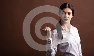 Portrait of serious brunette business woman pointing her thumb to the side in studio on brown background with copyspace.