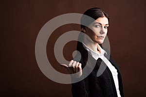 Portrait of serious brunette business woman pointing her thumb to the side in studio on brown background with copyspace.