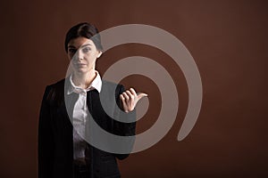 Portrait of serious brunette business woman pointing her thumb to the side in studio on brown background with copyspace.