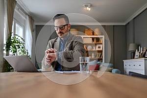 Portrait of serious businessman checking time on wristwatch while working over laptop in home office