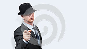 Portrait of serious businessman dressed in suit and hat pointing finger at camera, over light blue background.