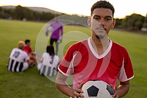 Portrait of serious caucasian male soccer player in red uniform holding ball at playground at sunset