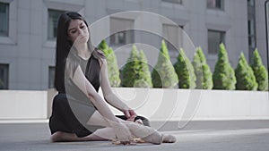 Portrait of serious charming woman tying up pointe shoes. Young confident Caucasian ballerina in black clothes sitting