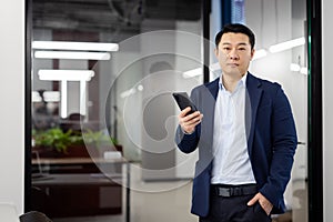 Portrait of serious concentrated businessman asian, man with phone in hands looking at camera, mature boss inside office