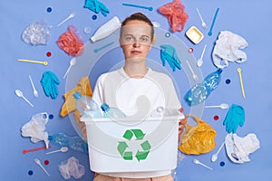 Portrait of serious concentrated girl with dark hair holding box of plastic bottles and recycling green symbol, sorting rubbish,