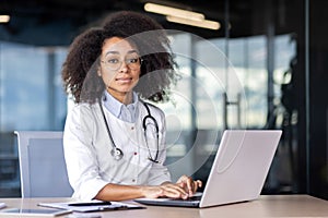 Portrait of a serious confident female doctor sitting in a hospital office at a desk, working on a laptop and looking at