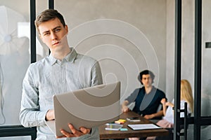 Portrait of serious confident young businessman in casual clothes holding laptop in hand standing at office looking at