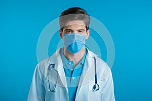 Portrait of serious doctor in professional medical mask and coat. Doc man isolated on blue background.