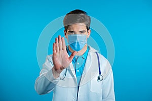 Portrait of serious doctor in professional medical white coat and mask showing rejecting gesture by stop palm sign. Doc