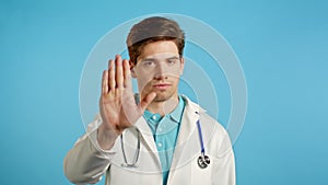 Portrait of serious doctor in professional medical white coat showing rejecting gesture by stop palm sign. Doc man