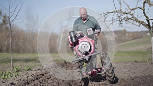 Portrait of serious elderly Caucasian man walking along rural garden with furrower. Old farmer furrowing fertile soil in