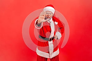 Portrait of serious elderly man with gray beard wearing santa claus costume showing stop ban gesture