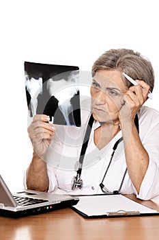 Portrait of serious elderly woman doctor sitting at table with computer and looking at x-ray