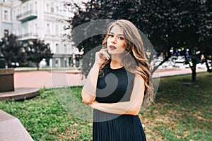 Portrait of serious elegante girl in the city park. She has long hair, black dress, vinous lips and looks wistfully