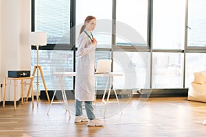 Portrait of serious female doctor in white coat standing on background of window in sunny day in light medical clinic