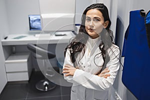 Portrait of serious female medic with arm crossed in CT scan room. Woman near the tomograph.