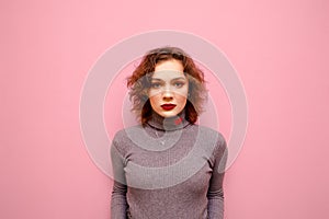 Portrait of a serious girl in a gray sweater stands on a pink background and looks into the camera with a serious face. Sad girl