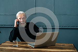 Portrait of serious gray-haired senior adult business man talking on mobile phone sitting at wooden table with laptop