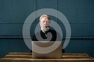 Portrait of serious gray-haired senior aged male sitting at wooden table at home office working using computer laptop