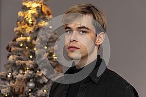 Portrait of a serious guy 20-25 years old in a black shirt on the background of a decorated Christmas tree.