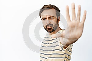 Portrait of serious guy extends his hand to stop you, showing palm taboo gesture, prohibit smth bad, standing over white