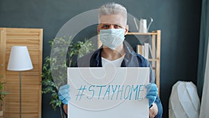 Portrait of serious guy in face mask and gloves holding stayhome banner in apartment